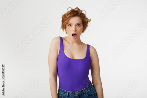 Young surprised attractive short-haired girl, wearing a purple jersey, looking at the camera in surprise with wide open mouth and eyes, she heard the shocking news. Isolated over white wall. © timtimphoto