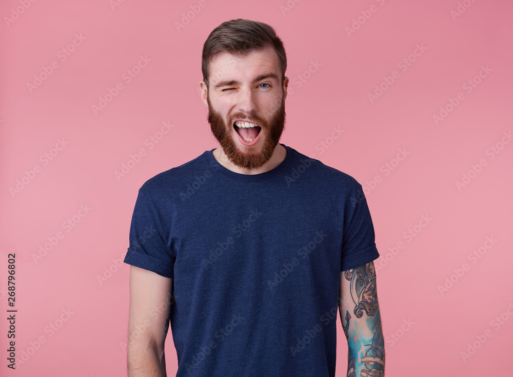 Φωτογραφία Young attractive red-bearded guy with blue eyes, wearing a blue t-shirt, looking at the camera and winking, look cool isolated over pink background