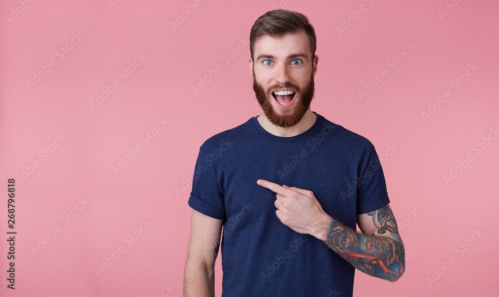 Φωτογραφία Young happy amazed attractive red-bearded young guy , wearing a blue t-shirt, with wide open mouth in surprise, pointing finger to copy space at the left side isolated over pink background