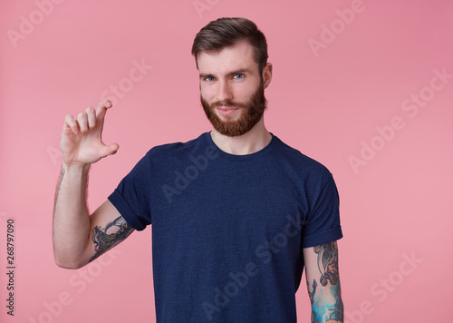 Obraz na plátně Portrait of young attractive red-bearded guy, frowning and looking at the camera, shows fingers something small with an insidious grin slyly looking at the camera isolated over pink background