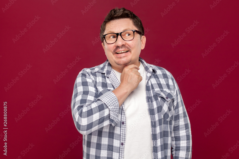 Fototapeta premium Portrait of smiley pondering handsome middle aged business man in casual checkered shirt and eyeglasses standing looking away and dreaming. indoor studio shot, isolated on dark red background.