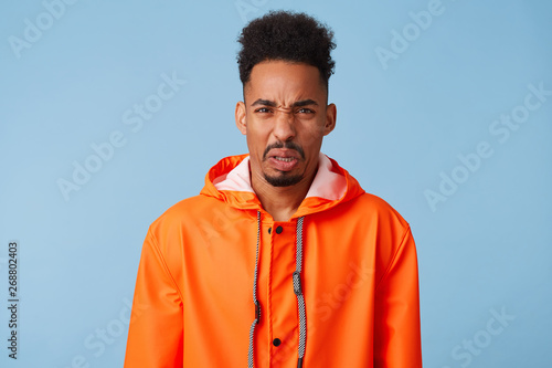 Close up of uhappy young african american dark skinned man in disgust, dissatisfied with the rainy weather outside, wears in orange rain coat, frowns isolated over blue background.