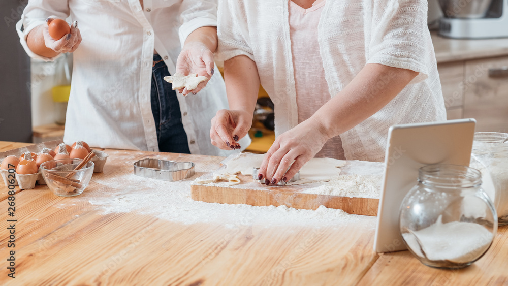 Midsection of two women making biscuit using cookie cutter to cut out dough. Home baking. Tablet with pastry recipe on. Midsection of two women making biscuit using cookie cutter to cut out dough. Home baking. Tablet with pastry recipe on.