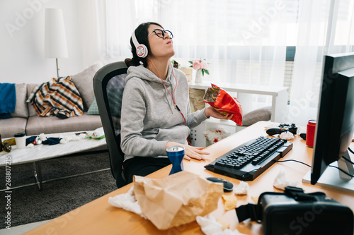 lazy college girl in messy dirty bedroom with order in junk food snack enjoy unhealthy life on summer break. happy young asian weird woman with headphones and glasses laughing binge watching online