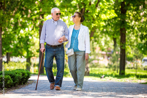 Cheerful senior couple having good time in city park, walking, laughing and enjoying sunny day. Old people wearing color clothes ans sunglasses