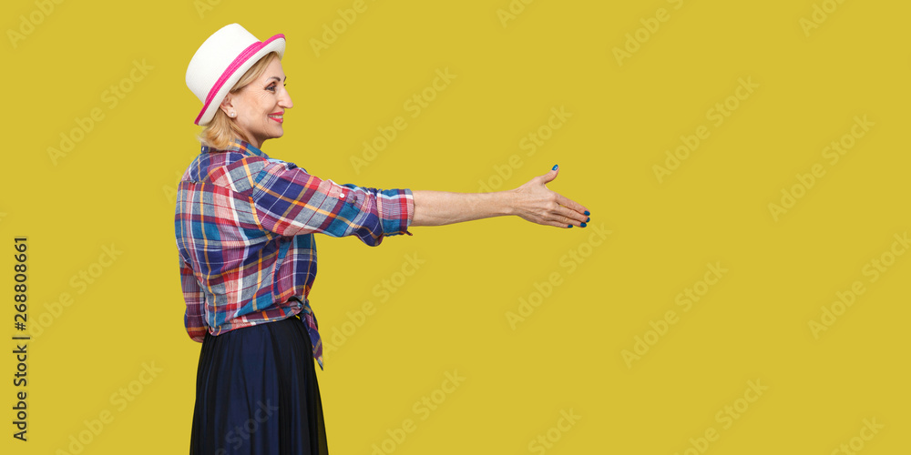 Profile side view portrait of happy modern stylish mature woman in casual style with hat standing, looking with toothy smile, giving hand to greeting. indoor studio shot isolated on yellow background.