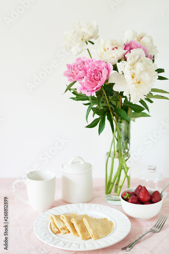 pancakes with strawberries and coffee on the table near a vase with peonies on a white background