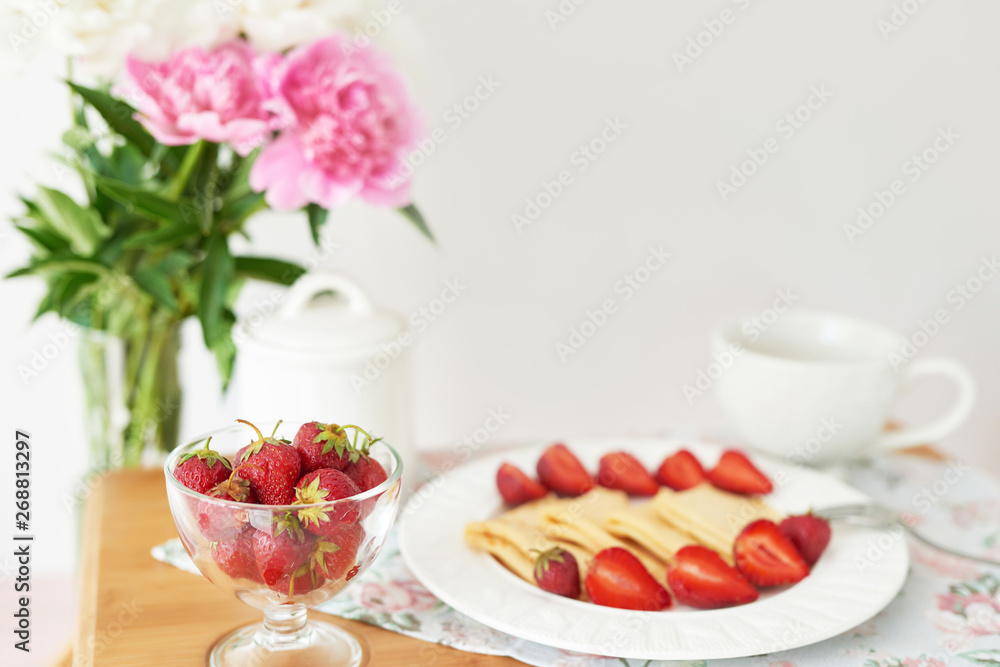pancakes with strawberries and coffee on the table near a vase with peonies on a white background
