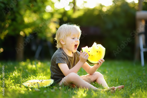 Caucasian little boy with blond hairs eating yellow watermelon on backyard