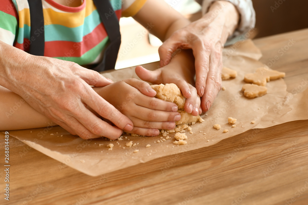 Little girl with her grandmother cooking cookies at table, closeup - obrazy, fototapety, plakaty 