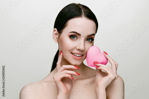 Portrait of model with natural nude make up. Beautiful young woman with perfect glow skin. Pink heart shaped konjac sponge. Organic beauty routine for massage, deep pore cleansing and exfoliating.