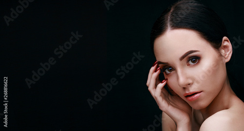 Portrait of brunette model with natural evening make up with bare shoulders on dark background. Beautiful young woman with brown eyes, long eyelashes and perfect glow clean skin touching her face. 
