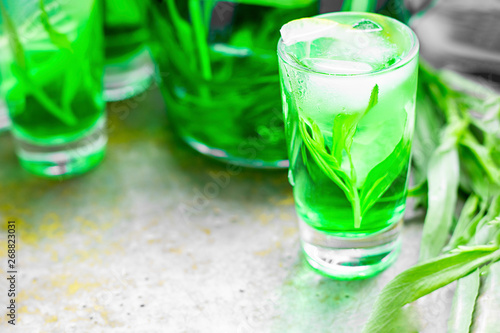 Summer cold green drink with tarragon, mint and lemon in small shot with ice and defocused decanter on background. Sparkling beverage, alcohol drink on dark background. Horizontal, copy space