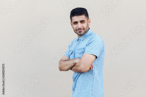 Portrait of confident successful handsome young bearded businessman in blue shirt standing, crossed arms and looking at camera with smile. indoor studio shot isolated on light beige wall background.