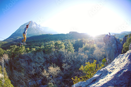A man is walking along a stretched sling. Highline in the mountains. Man catches balance. Performance of a tightrope walker in nature. Highliner on the background of valley. photo