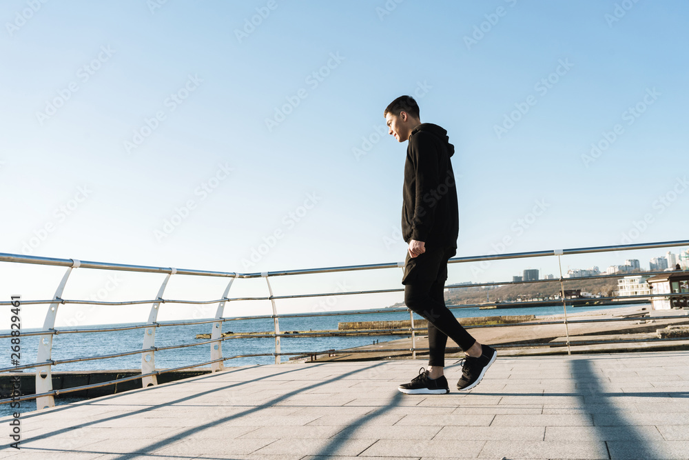 Photo of joyful guy 20s walking along wooden boardwalk by seaside after morning workout