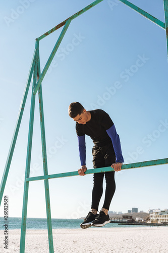 Photo of active guy doing acrobatics on horizontal gymnastic bar during morning workout by seaside