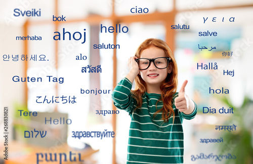 education, translation and learning concept - smiling red haired student girl in glasses and green striped shirt over greeting words in different foreign languages on background photo