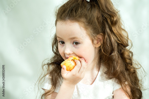 Cute little girl nibbles an apple with pleasure. Sun glare on the face. Emotional portrait of a little beautiful girl holding an apple. Studio 4-5 years.