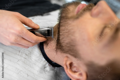 grooming and people concept - man and barber with trimmer or shaver cutting beard at barbershop