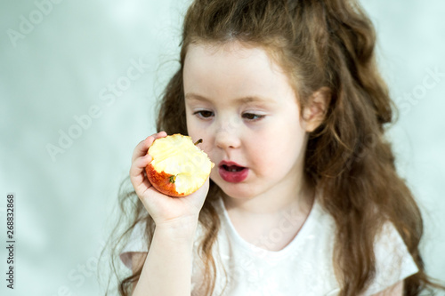 Cute little girl nibbles an apple with pleasure. Sun glare on the face. Emotional portrait of a little beautiful girl holding an apple. Studio 4-5 years.