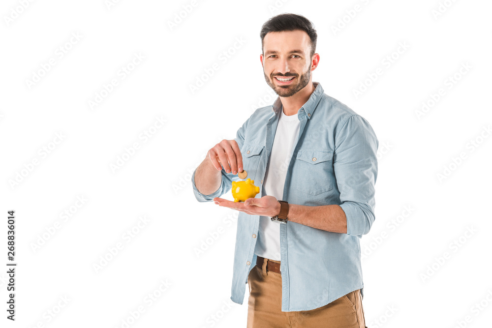 cheerful bearded man putting coin into piggy bank and looking at camera isolated on white cheerful bearded man putting coin into piggy bank and looking at camera isolated on white