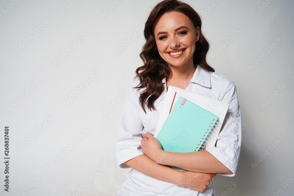 Beautiful smiling young female doctor in white medical jacket isolated on white background. Brunette woman cosmetologist holding books, notebook, folder. Copy space Beautiful smiling young female doctor in white medical jacket isolated on white background. Brunette woman cosmetologist holding books, notebook, folder. Copy space