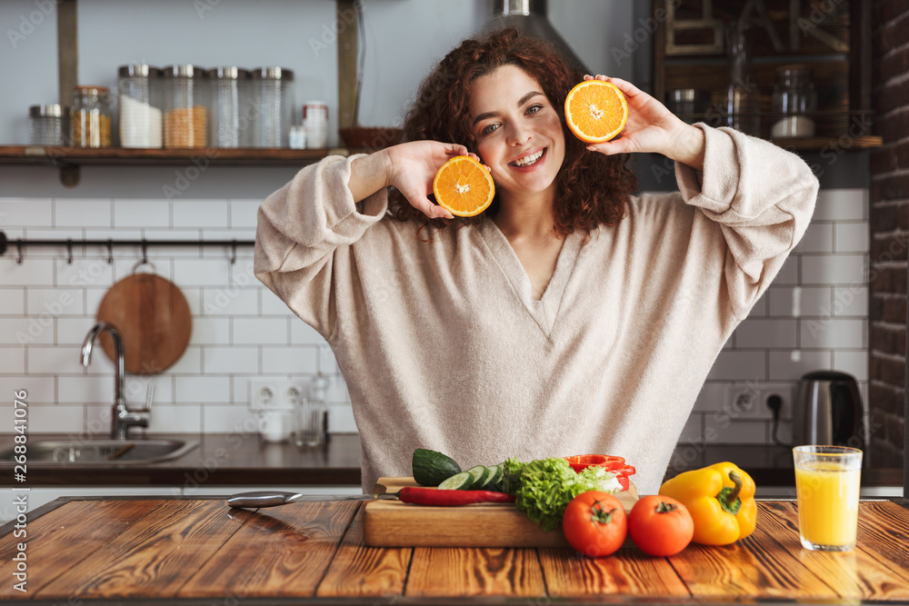 Photo of cute caucasian woman smiling and holding two orange parts in kitchen interior at home