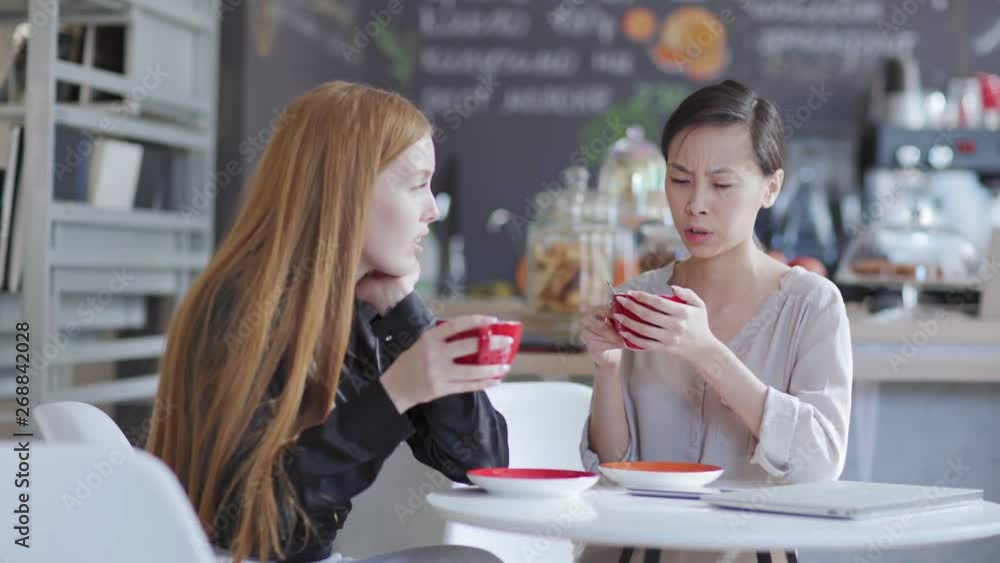 Tracking right shot of two young women sitting at table in cafe, having coffee and talking. Asian female telling story emotionally to red-haired Caucasian friend Tracking right shot of two young women sitting at table in cafe, having coffee and talking. Asian female telling story emotionally to red-haired Caucasian friend
