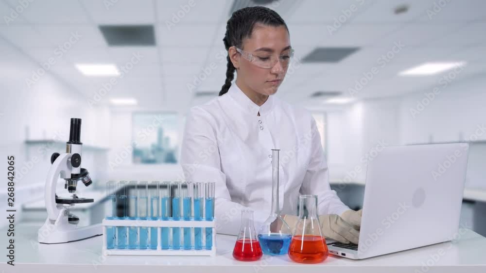 A female scientist writes the results of a genetic study to a laptop while sitting at a white table in a chemical laboratory. A female scientist writes the results of a genetic study to a laptop while sitting at a white table in a chemical laboratory.
