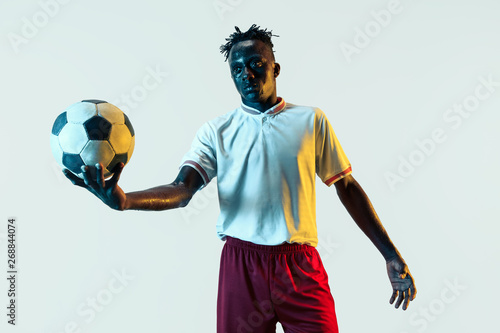 Young african-american male football or soccer player in sportwear and boots standing with the ball in neon light isolated on white background. Concept of healthy lifestyle, professional sport, hobby. © master1305