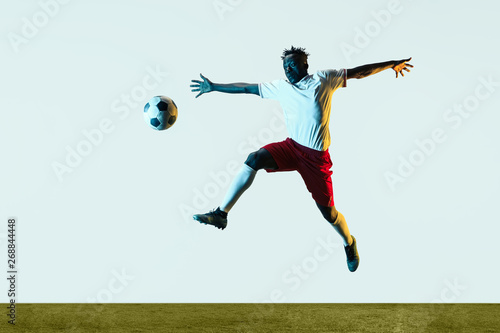 Young african-american male football or soccer player in sportwear and boots kicking ball for the goal in neon light isolated on white background. Concept of healthy lifestyle, professional sport