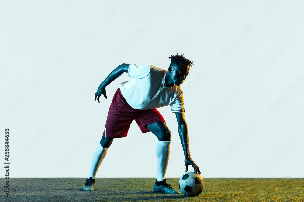 Young african-american male football or soccer player in sportwear and boots kicking ball for the goal in neon light isolated on white background. Concept of healthy lifestyle, professional sport