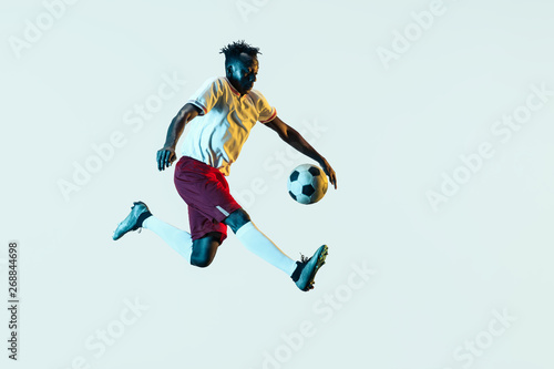 Young african-american male football or soccer player in sportwear and boots kicking ball for the goal in neon light isolated on white background. Concept of healthy lifestyle, professional sport