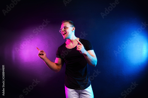 Excited young male in trendy outfit pointing away while having fun under colorful illumination during party