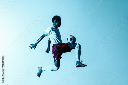 Young african-american male football or soccer player in sportwear and boots kicking ball for the goal in jump in neon light on gradient background. Concept of healthy lifestyle, professional sport.