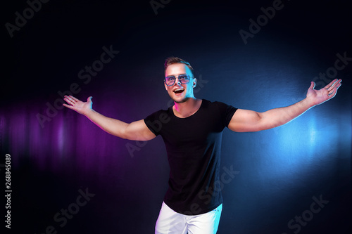 Cheerful young guy in stylish outfit stretching out arms and looking at camera while standing under colorful light during party