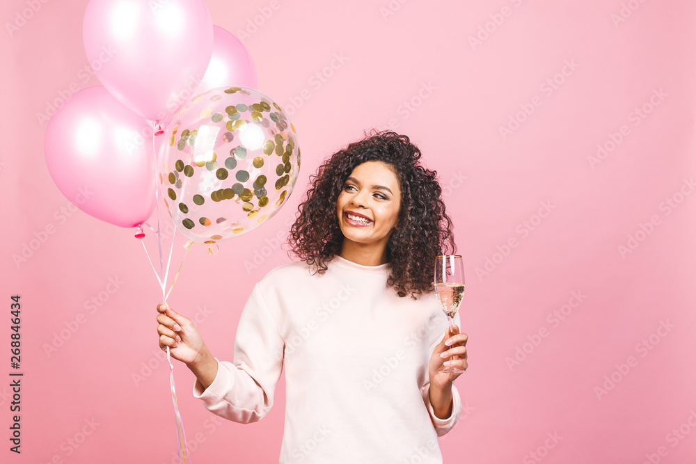 Party time! Happy beautiful african-american woman with glass of champagne, balloons and falling confetti isolated over pink background. - obrazy, fototapety, plakaty 