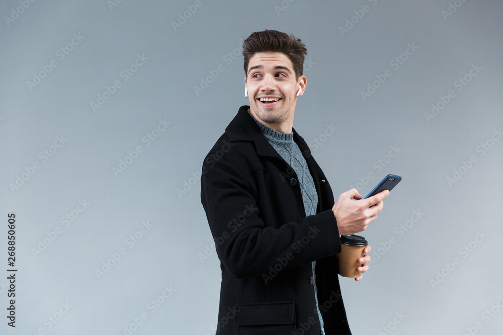 Image of joyful young man wearing earpods holding takeaway coffee and mobile phone Image of joyful young man wearing earpods holding takeaway coffee and mobile phone