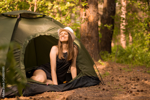 Happy woman in hat sits in tent around forest under sunset light sky enjoying leisure and freedom.