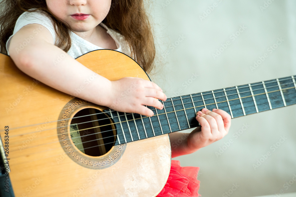 Fototapeta premium Portrait of a little girl with a guitar. For the first time the girl was given a guitar. Nearby sits a teddy bear. A child of 5 years old is studying a musical instrument.