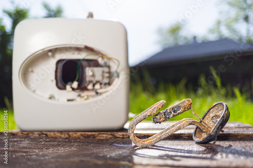 The water heating element damaged from corrosion lies on a wooden table next to the boiler. In the background green grass in the yard photo