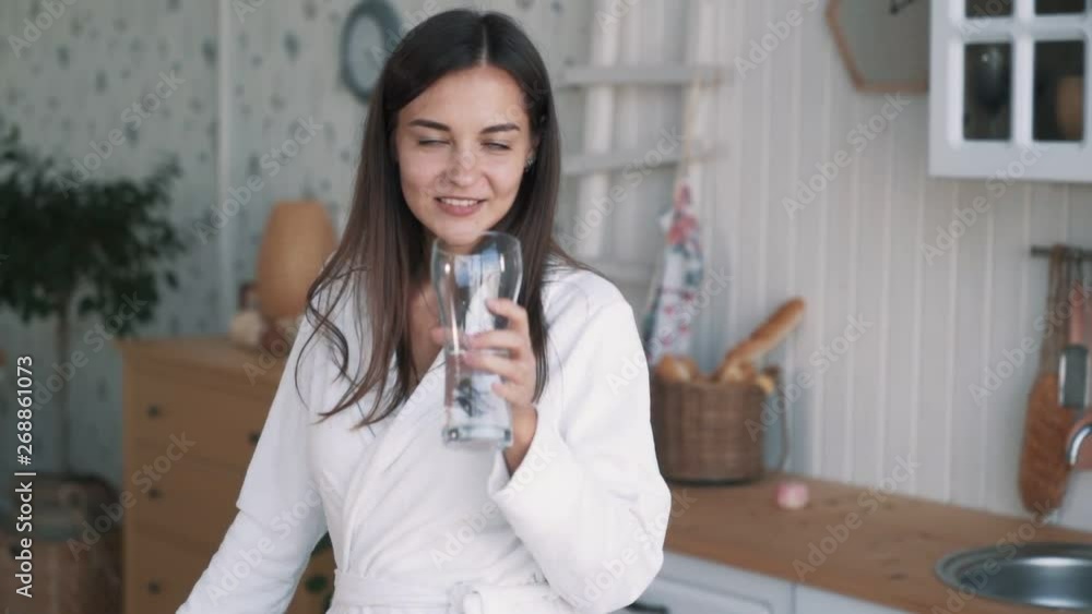 Girl in bathrobe looks at camera, smiles, drinks water from glass, slow motion