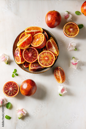Group of fresh organic Sicilian blood oranges sliced and whole in ceramic plate, edible flowers, mint leaves over white marble background. Flat lay, space