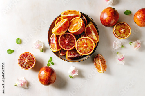Group of fresh organic Sicilian blood oranges sliced and whole in ceramic plate, edible flowers, mint leaves over white marble background. Flat lay, space