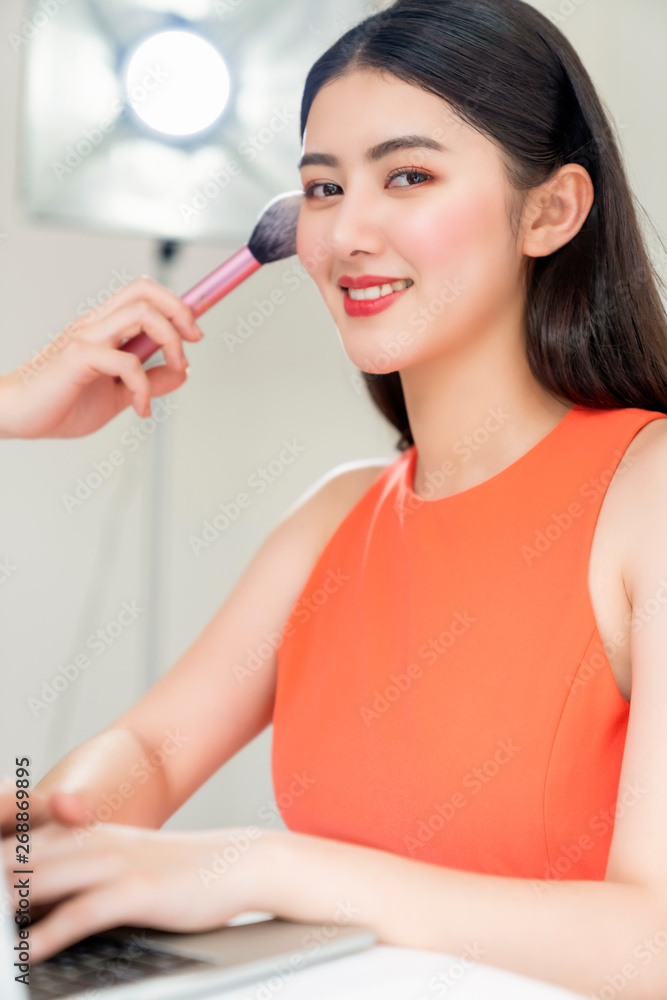 asian pretty Pleasant cheerful professional TV announcer sitting at the table with laptop with the assistant making makeup with brush on asian pretty Pleasant cheerful professional TV announcer sitting at the table with laptop with the assistant making makeup with brush on