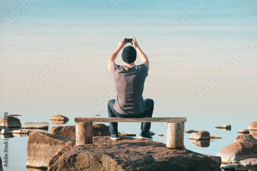 Fototapeta A man sits on a wooden bench and photographs the sea on his mobile phone