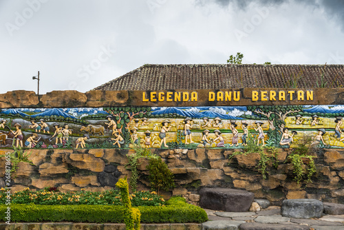 Bali, Indonesia - February 25, 2019: Ulun Danu Beratan Temple in Bedoegoel. In park, Colorful fresco depicting legend of region, set in brown stone wall with green foliage up front under cloudscape. photo