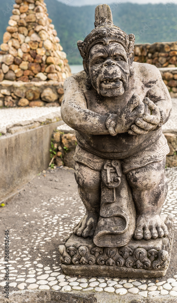 Bali, Indonesia - February 25, 2019: Ulun Danu Beratan Temple complex in Bedoegoel. Gray stone statue of dwarf in park.