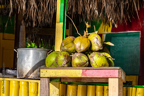 Tropical jelly coconut fruits on table at outdoor vendor shop painted in rasta colors with thatch roof. Big pot of Jamaican soup dish cooking in background. Sunny summer beach day setting in Jamaica. photo